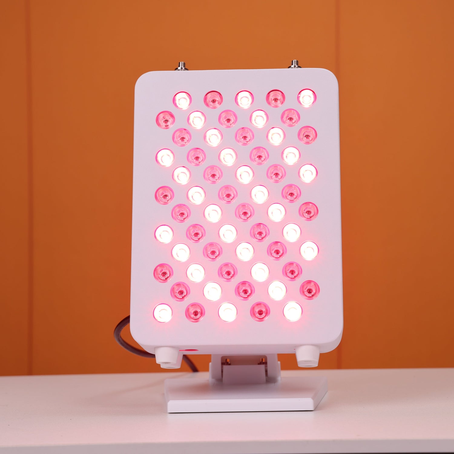 Front view of a Nicebeam red light therapy panel showcasing its array of 60 LED lights evenly distributed, glowing with a therapeutic red light, mounted on a stable stand, and set against a vibrant orange background.